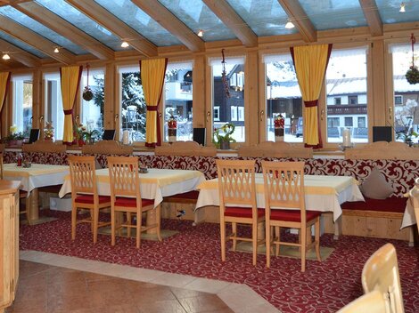 Ein gemütlicher Restaurantbereich mit Holzstühlen und Tischen. Große Fenster lassen viel Licht herein und bieten einen Blick nach draußen.