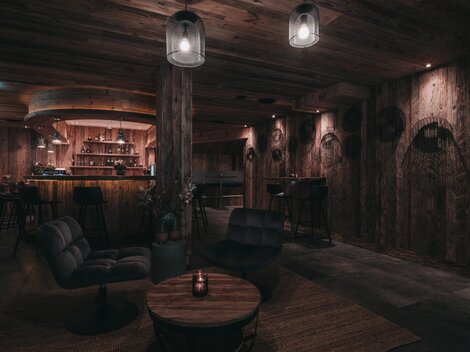 Ein gemütlicher Raum mit Holzwänden und sanfter Beleuchtung. Die Bar im Hintergrund und die modernen Möbel schaffen eine einladende Atmosphäre.