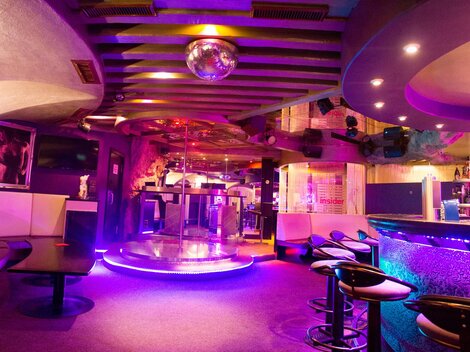 Ein stilvoller Club mit einem beleuchteten Barbereich und einer Tanzfläche in der Mitte. Die bunten Lichter schaffen eine lebhafte Atmosphäre.