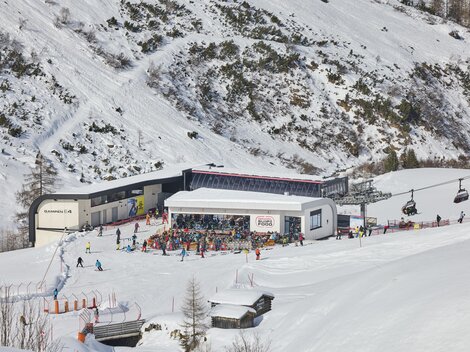Eine belebte Skistation in den Bergen mit vielen Skifahrern. Im Hintergrund sind schneebedeckte Hänge und eine Seilbahn zu sehen.