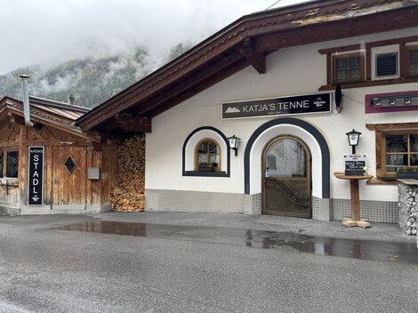 Ein traditionelles alpenländisches Gebäude mit einem einladenden Eingang. Neben dem Gebäude befindet sich ein Holzstapel und ein Schild mit der Aufschrift "STADL".