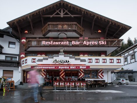 Ein gemütliches Restaurant mit einer Bar im Après-Ski-Stil. Die Fassade ist rustikal mit Holzdetails und einem einladenden Außenbereich.
