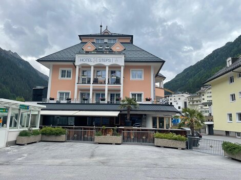 Ein charmantes Hotel mit einer hellen Fassade und Balkon im Erdgeschoss. Im Hintergrund sind grüne Berge und eine ruhige Atmosphäre zu sehen.