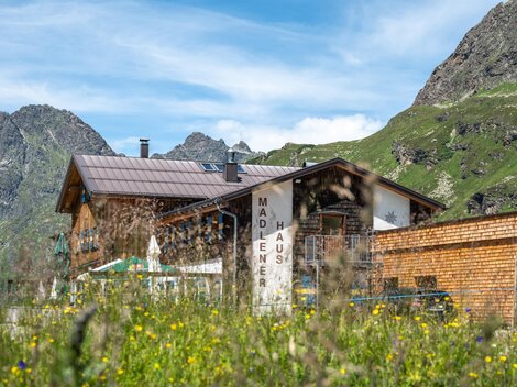 Ein rustikales Haus umgeben von grünen Bergen und bunten Wiesenblumen. Im Vordergrund sind blühende Wiesen und im Hintergrund sind majestätische Berge sichtbar.