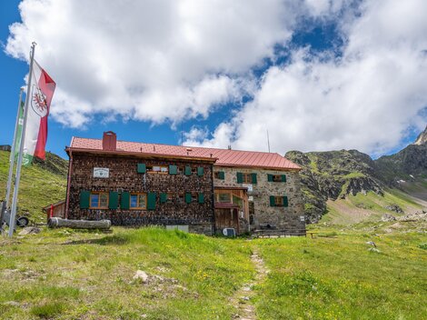 Eine Berghütte aus Stein inmitten einer grünen Wiese. Im Hintergrund sind Berge und ein blauer Himmel mit einigen Wolken zu sehen.