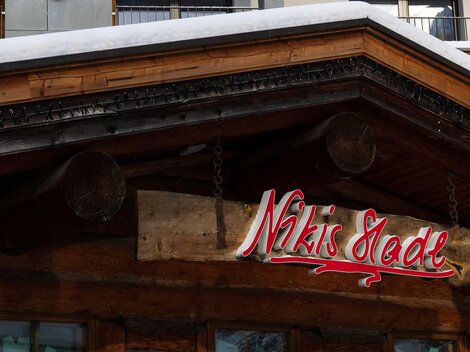 Ein traditionelles Holzgebäude mit einem schneebedeckten Dach. Das Schild mit der Aufschrift "Nikis Stube" ist gut sichtbar.