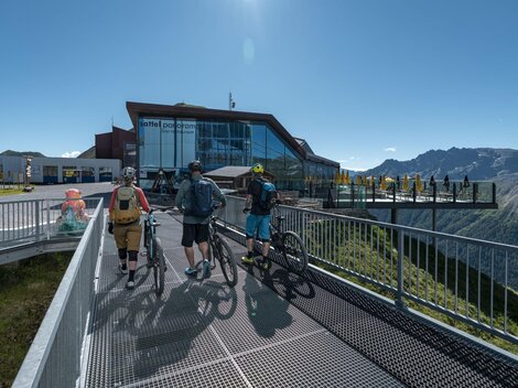 Drei Radfahrer stehen auf einer Aussichtsplattform mit Blick auf die Berge. Im Hintergrund ist ein modernes Gebäude zu sehen.