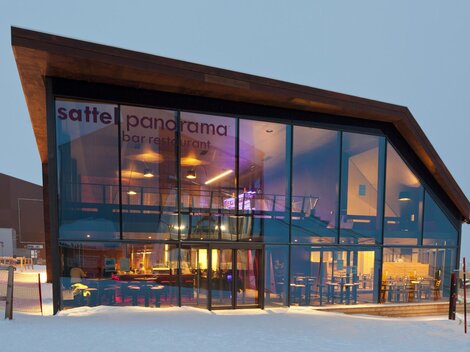 Ein modernes Restaurant mit großen Glasfenstern und einem schlichten Holzdesign. Das Gebäude liegt in einer winterlichen Umgebung mit Schnee.