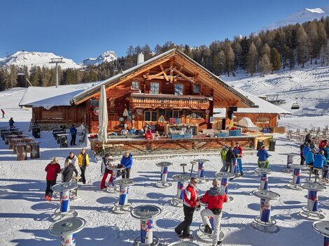 Eine gemütliche Berghütte in der verschneiten Landschaft, umgeben von Bäumen. Viele Menschen genießen die winterliche Atmosphäre und stehen an Tischen im Freien.
