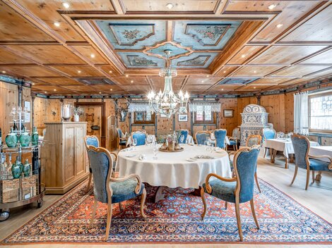 Ein eleganter Restaurantbereich mit einem runden Tisch und blauen Stühlen. Das Zimmer ist mit Holzverkleidungen, einem kunstvollen Deckenmuster und einem prächtigen Kronleuchter dekoriert.