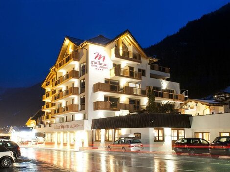 Ein modernes Hotelgebäude mit mehreren Stockwerken und Balkonen. Die Umgebung ist abends beleuchtet, und die Straßen sind nass vom Regen.