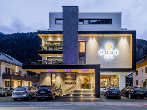 Ein modernes Hotelgebäude mit dem Schriftzug „POST“ leuchtet in der Dämmerung. Umgeben von Bergen und anderen Gebäuden ist es ein einladender Ort zum Übernachten.