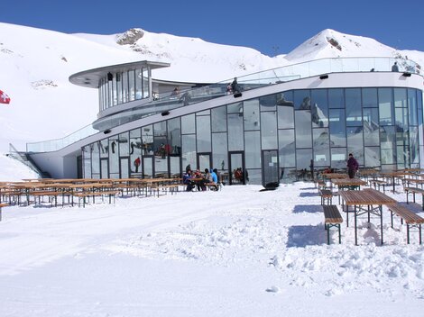 Ein modernes Gebäude in den Bergen, umgeben von Schnee. Draußen stehen Holzbankreihen und Menschen genießen die winterliche Landschaft.
