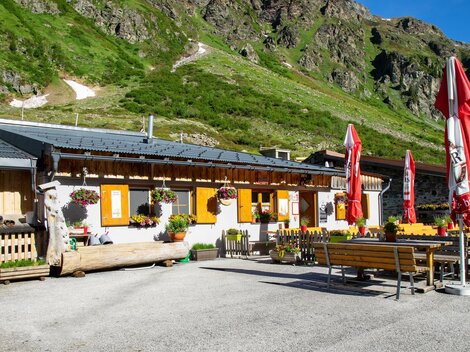 Eine gemütliche Almhütte mit bunten Blumen und roten Schirmen. Im Hintergrund sind grüne Berge zu sehen.