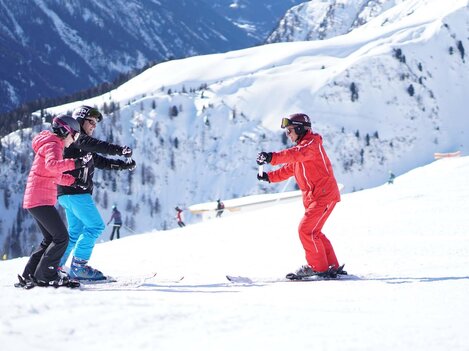 Drei Personen beim Skiunterricht auf einer verschneiten Piste. Im Hintergrund sind beeindruckende Berge zu sehen.