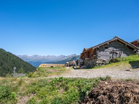 Ein malerisches Bergdorf mit traditionellen Holzgebäuden und einer wunderschönen Aussicht auf die Berge. Der strahlend blaue Himmel und die grüne Wiese verleihen der Szene eine serene Atmosphäre.