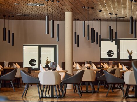 Ein modernes Restaurant mit eleganten Tischen und Stühlen. Die Decke ist mit schlichten Hängeleuchten dekoriert und große Fenster sorgen für viel Licht.