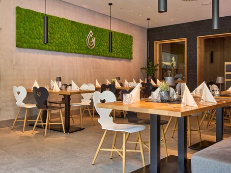 Ein modernes Restaurant mit stilvollen Tischen und Stühlen. An der Wand ist eine lebendige Pflanzenwand zu sehen, die eine einladende Atmosphäre schafft.