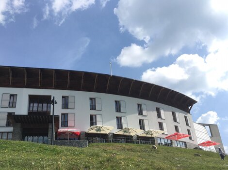 Ein modernes Gebäude in den Bergen, umgeben von Wiesen und blühenden Pflanzen. Der Himmel ist klar und blau.
