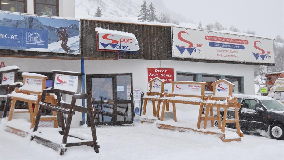 Ein Sportgeschäft im Winter mit viel Schnee vor der Tür. Die Fassade zeigt Schilder und Holzgestelle für Skiausrüstung.
