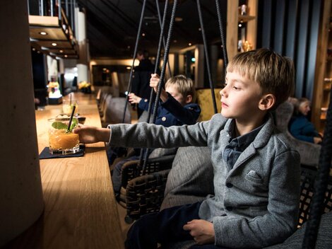 Ein Junge in einem schlichten grauen Anzug sitzt an der Bar und greift nach einem Getränk. Im Hintergrund sind weitere Kinder und eine gemütliche Innenausstattung sichtbar.