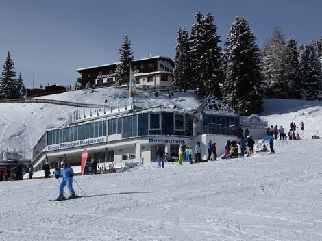 Ein schneesicherer Skikurs mit einem modernen Restaurant im Vordergrund. Viele Menschen genießen aktiv den Winterspaß in einer malerischen Berglandschaft.