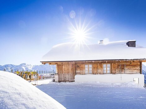 Ein charmantes Holzhaus im Schnee mit strahlender Sonneneinstrahlung. Im Hintergrund sind schneebedeckte Berge sichtbar.