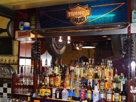 Eine Bar mit dem Namen "Tommy's Garage", voll mit verschiedenen Alkoholflaschen und Gläsern. An der Wand hängen Reifen und die Beleuchtung ist gemütlich.