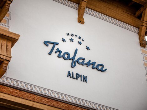 Ein Hotel-Schild mit dem Namen "Trofana Alpin" ist an der Wand angebracht. Die Fassade zeigt rustikale Holzelemente und eine elegante Schriftart.