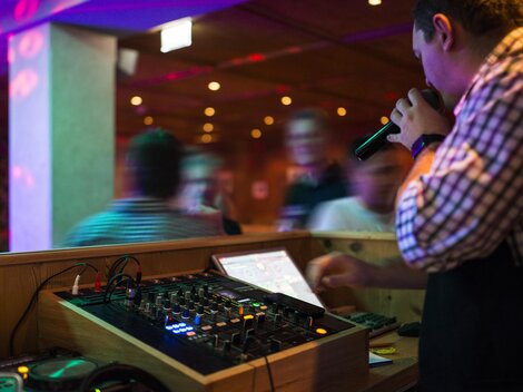 Ein DJ steht hinter einem Mischpult und hält ein Mikrofon in der Hand. Die bunten Lichter und die tanzende Menge schaffen eine lebendige Partystimmung.