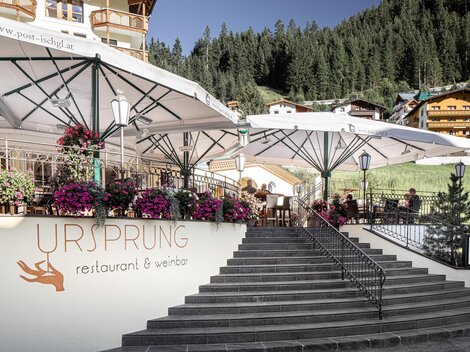 Ein gemütliches Restaurant mit Terrassen und blühenden Blumen. Im Hintergrund sind grüne Berge und traditionelle Hütten zu sehen.