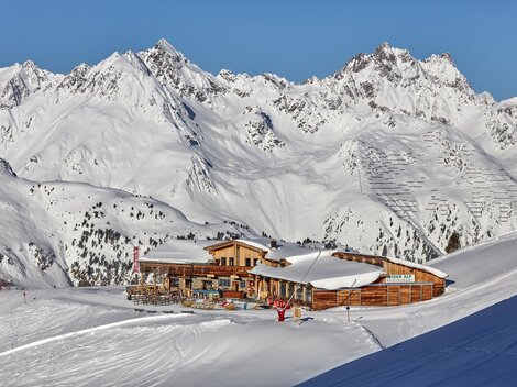 Eine gemütliche Berghütte liegt in einer schneebedeckten Landschaft. Im Hintergrund ragen majestätische Berge unter einem klaren Himmel empor.