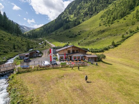 Eine malerische Landschaft mit einem Gasthaus in den Bergen. Fluss und grüne Wiesen umgeben das Gebäude.