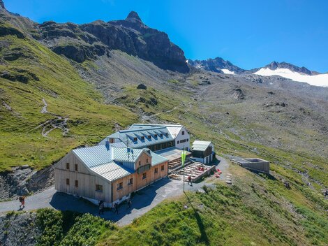 Eine Berghütte umgeben von grünen Wiesen und Bergen. Der Himmel ist klar und blau, ideal für einen Outdoor-Urlaub.