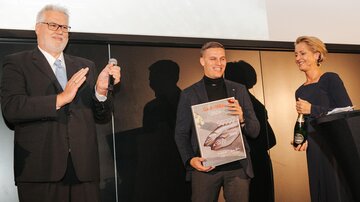 Eine Preisverleihung mit drei Personen auf der Bühne. Der Preisträger hält eine Auszeichnung und lächelt, während die anderen applaudieren.